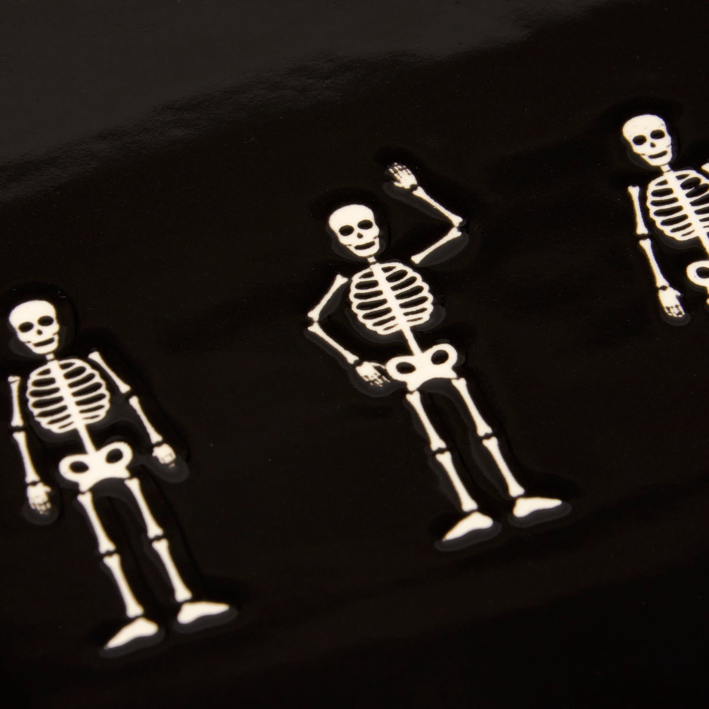 White skeleton-shaped enamel pins with black outlines in playful poses on a dark background for skeletons oval platter