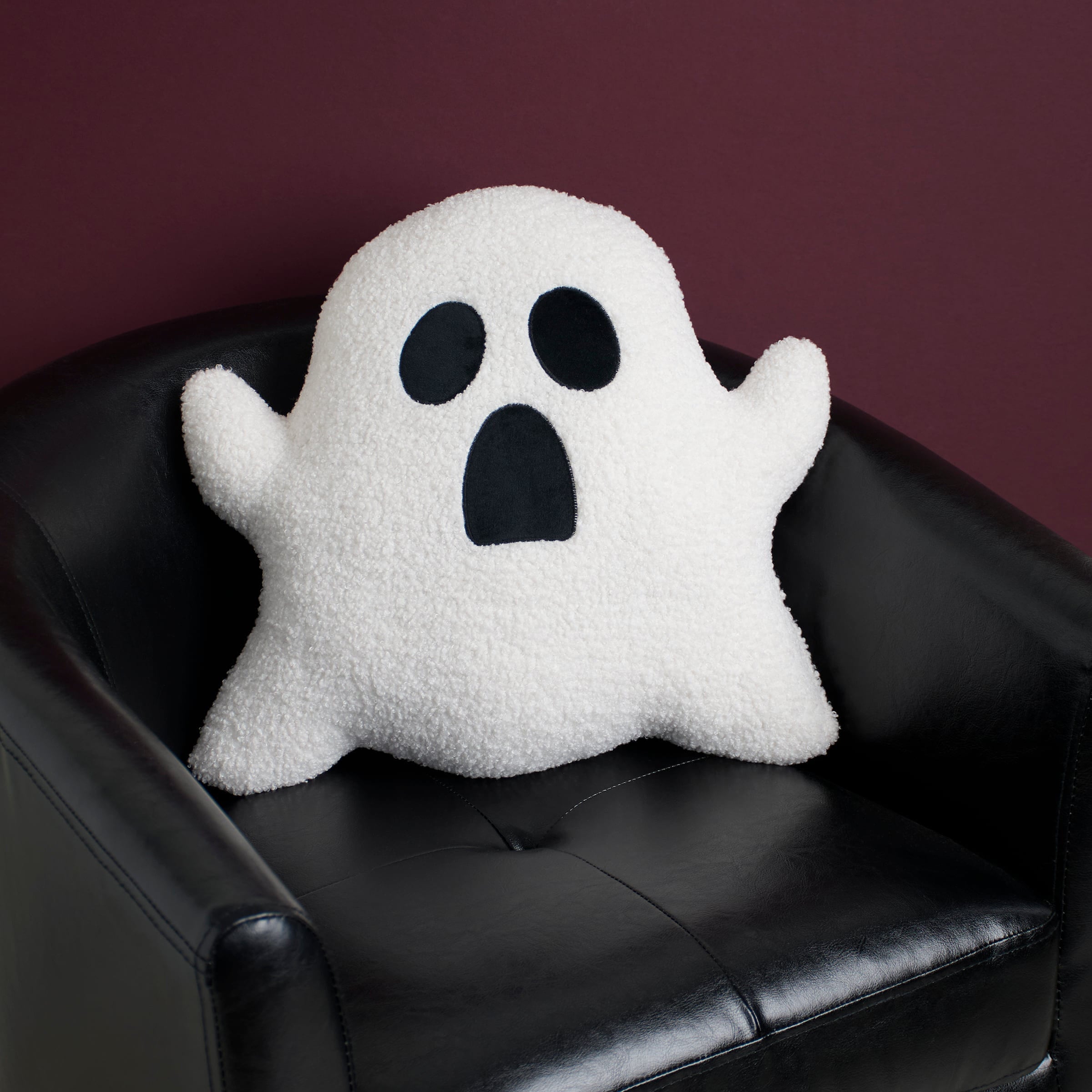 White plush ghost shaped pillow with black embroidered eyes and mouth sitting on a black leather chair