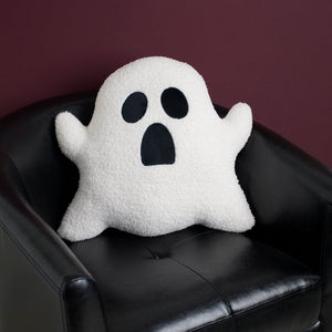 White plush ghost shaped pillow with black embroidered eyes and mouth sitting on a black leather chair