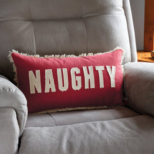 A red rectangular naughty nice pillow with frayed edges by dan dipaolo on a gray armchair perfect for holiday pillows