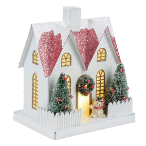 Lighted fenced house sitter with a white christmas village house, glittery red roof, lit windows, and mini flocked trees with red ornaments