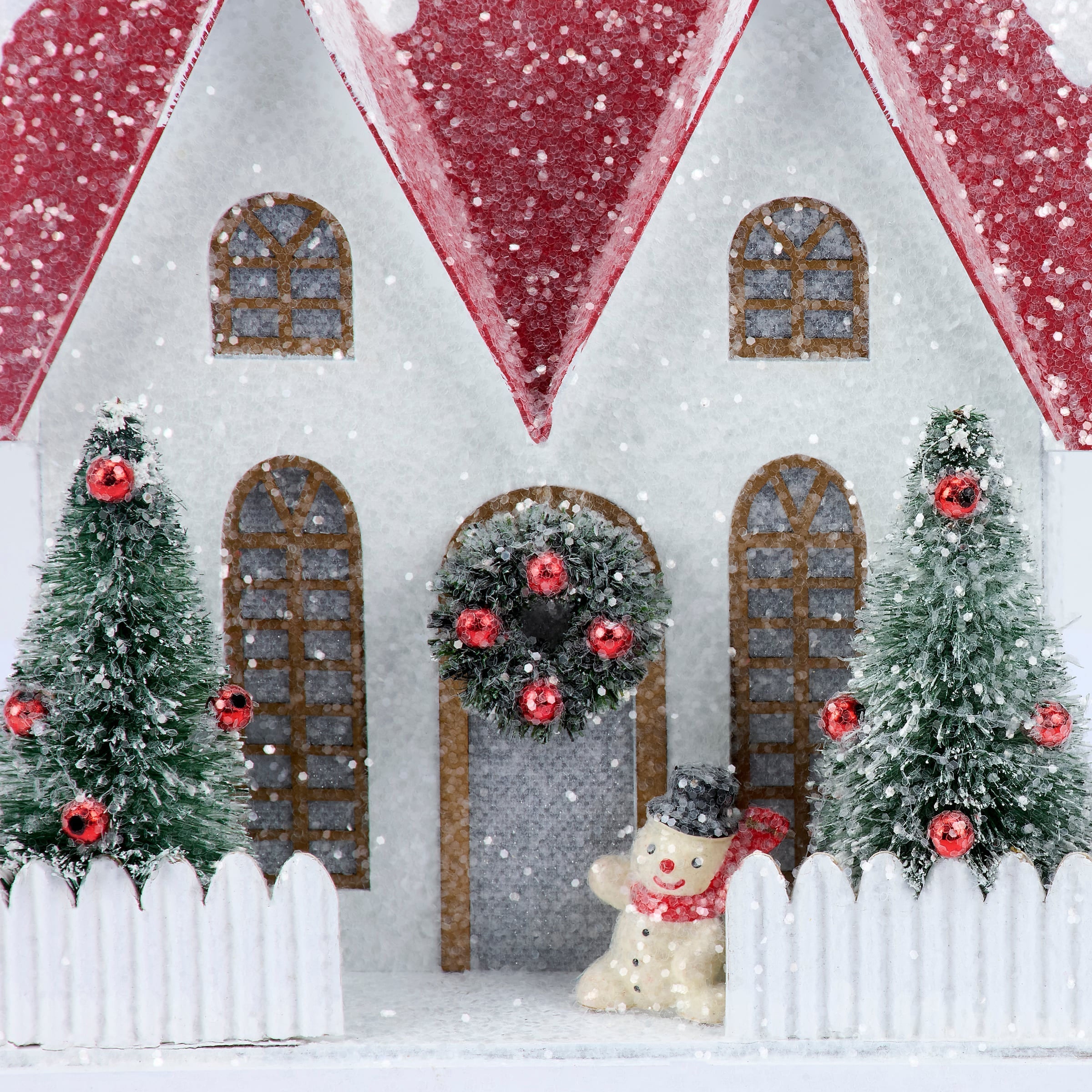 Lighted fenced house sitter with a white miniature house, red glittery roofs, and frosted green christmas trees with red ornaments