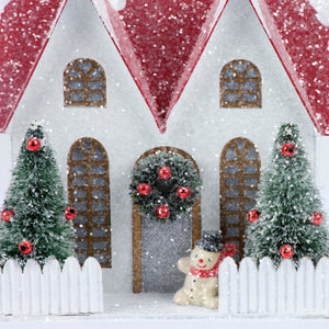 Lighted fenced house sitter with a white miniature house, red glittery roofs, and frosted green christmas trees with red ornaments
