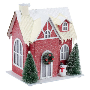A festive lighted red house sitter with glittery red exterior snow-dusted roof and miniature pine trees and snowman