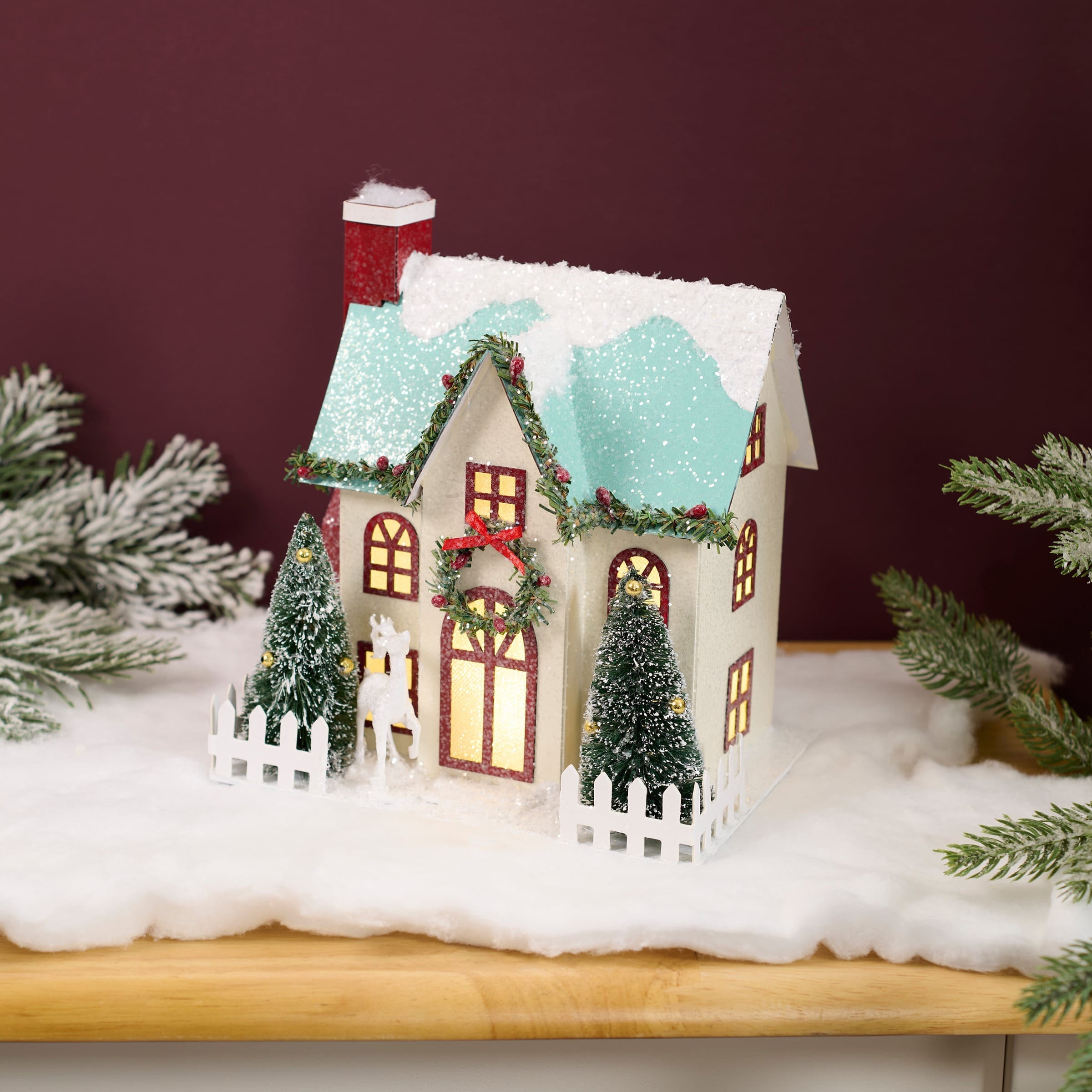 A charming white lighted wreath house sitter with red chimney, snow-dusted roof, and green garlands with berries