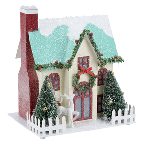 A festive lighted wreath house sitter with cream walls, teal snowy roof, red glitter chimney, and green garlands with red bows