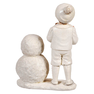 White glittery resin winter white kid building snowman figurine with boy in winter outfit standing beside snowman figure