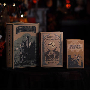Three vintage-style Halloween book props displayed on a dark table against a blurred warm background. The books read "The Book of Halloween," "Witches Potions & Spells," and "Creatures of the Night," with illustrated spooky scenes on tan and dark blue covers.