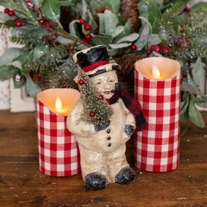 Display shot of a holiday snowman figurine stands on a wooden surface between two red-and-white checkered pillar candles, with green evergreen branches and red berries in the background.