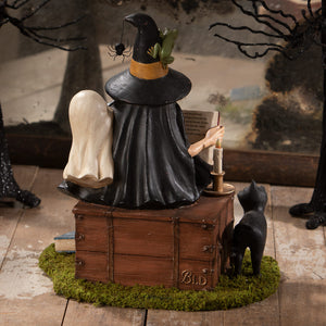 A black witch figurine with spider hat reading a book on a wooden chest candle halloween witches decor ghost stories