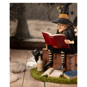 A detailed ghost stories witch figurine with glasses in black hat and cloak sitting on a chest reading a red book perfect for halloween witches and witch decor