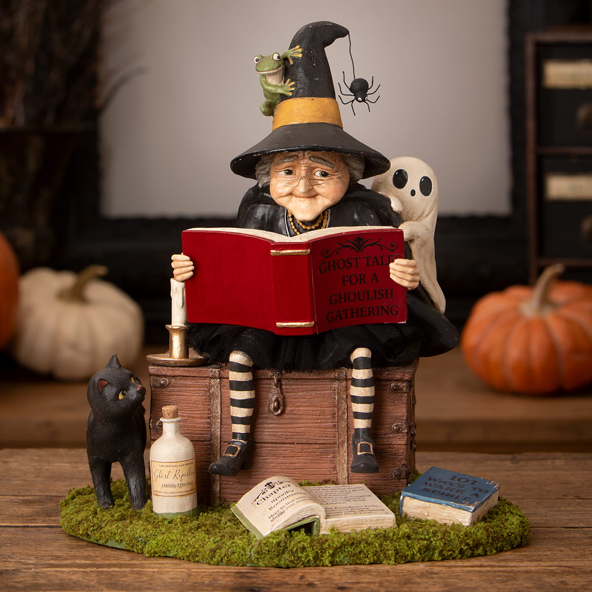 A whimsical ghost stories witch figurine with black and yellow hat reading a red book surrounded by halloween witches decor