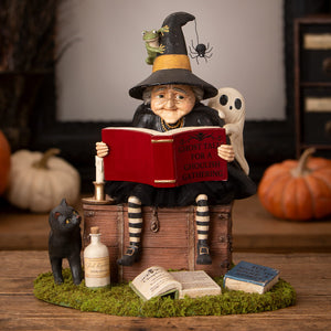 A whimsical ghost stories witch figurine with black and yellow hat reading a red book surrounded by halloween witches decor