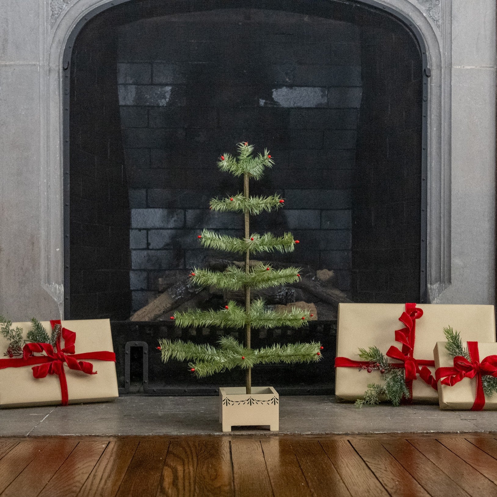 A small artificial Christmas tree stands centered on a fireplace hearth, flanked by wrapped gift boxes with red ribbons and evergreen sprigs. The fireplace has a dark brick interior and gray stone surround, with a wooden floor in front.
