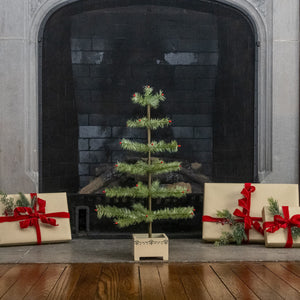 A small artificial Christmas tree stands centered on a fireplace hearth, flanked by wrapped gift boxes with red ribbons and evergreen sprigs. The fireplace has a dark brick interior and gray stone surround, with a wooden floor in front.