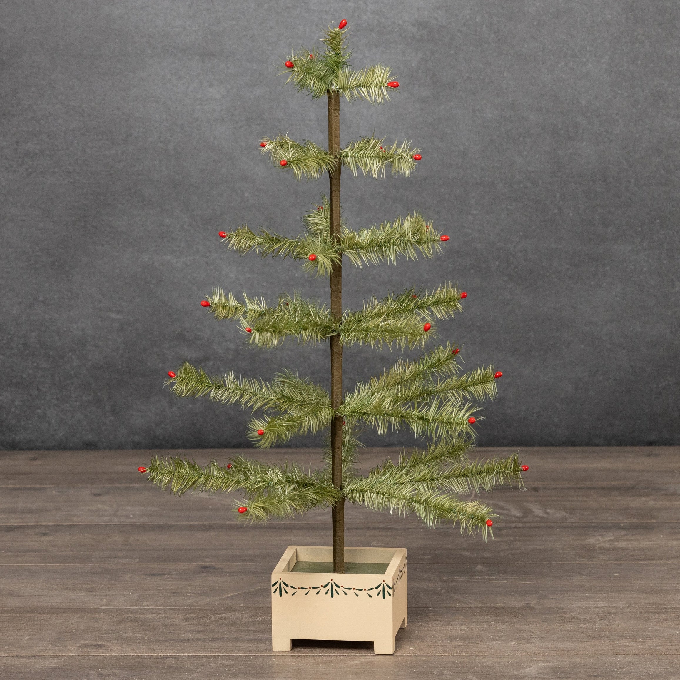 A small feather Christmas tree with green needles and red berries, mounted on a light wood base, set against a dark gray textured wall and wooden floor.