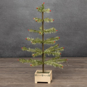 A small feather Christmas tree with green needles and red berries, mounted on a light wood base, set against a dark gray textured wall and wooden floor.
