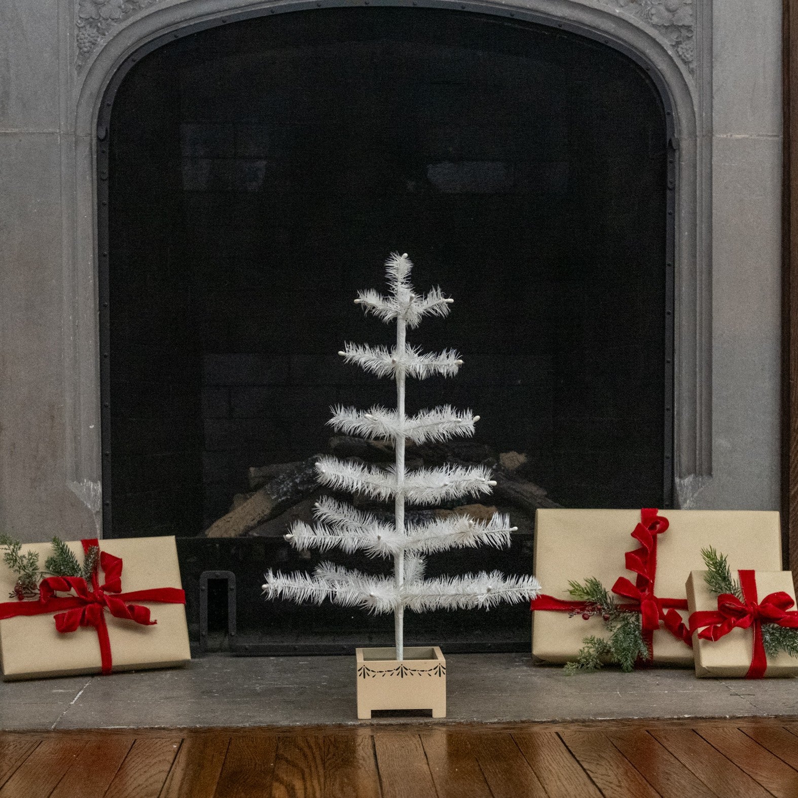 A small white artificial Christmas tree in a wooden base stands on a fireplace hearth, flanked by wrapped gift boxes with red ribbons and evergreen sprigs.