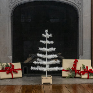 A small white artificial Christmas tree in a wooden base stands on a fireplace hearth, flanked by wrapped gift boxes with red ribbons and evergreen sprigs.