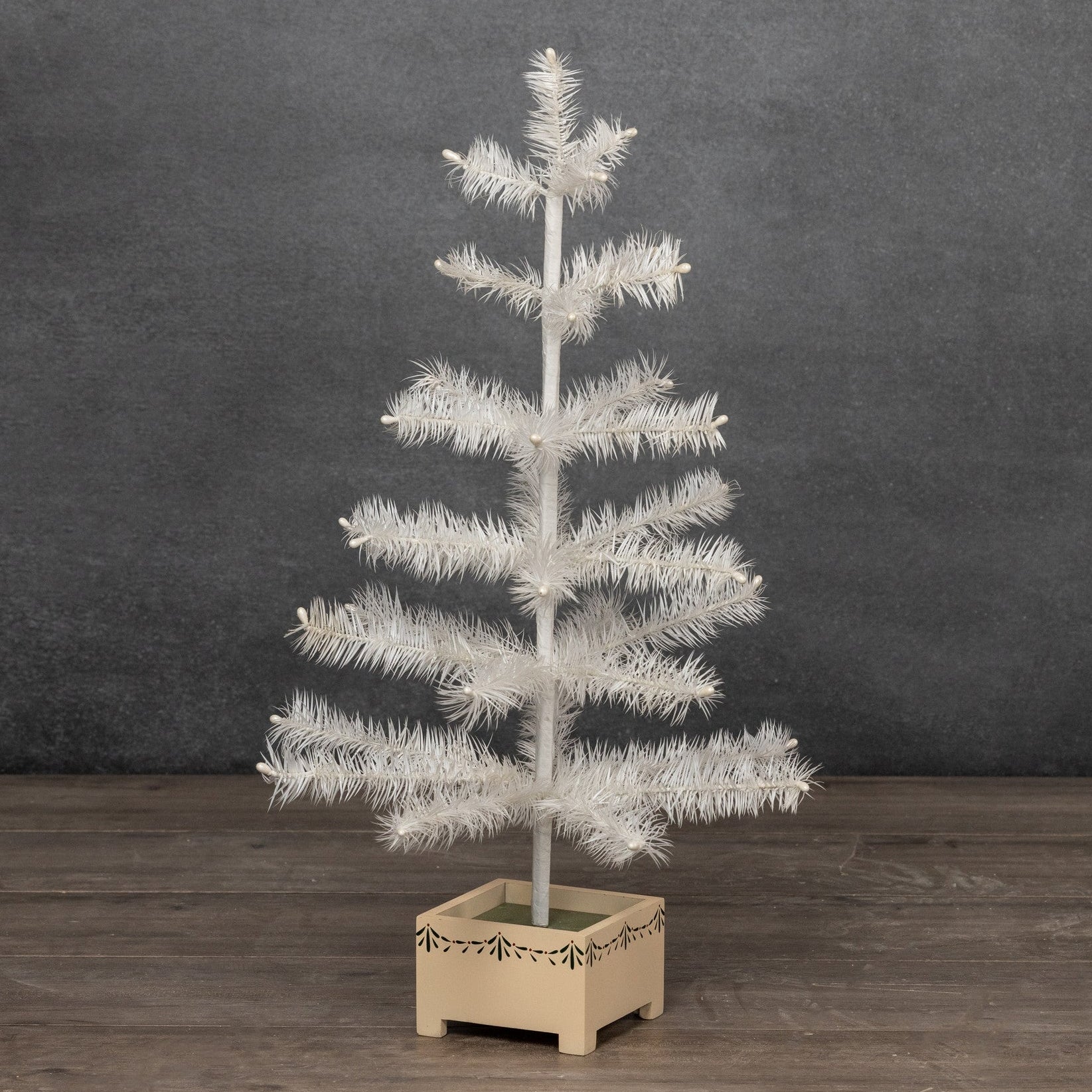 A small white feather Christmas tree with textured faux pine needles, standing in a light beige square wooden planter box on a dark wood floor against a dark gray textured wall.