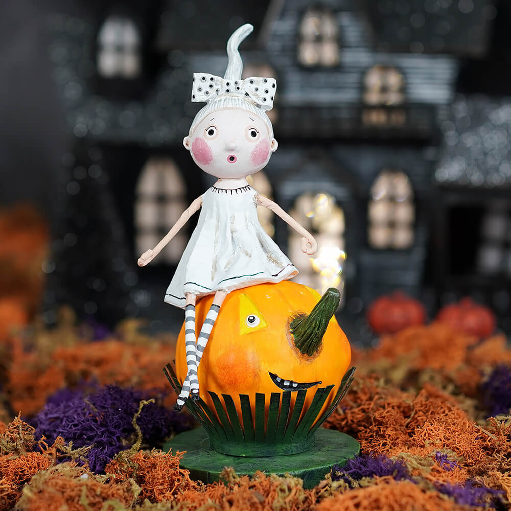 A whimsical lori mitchell halloween figurine with pink cheeks and bow sits on an orange jack o lantern for trick or treat kids