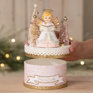 A delicate white and pink bethany lowe angel figure with gold wings holding a book atop glittery trees on peace on earth box