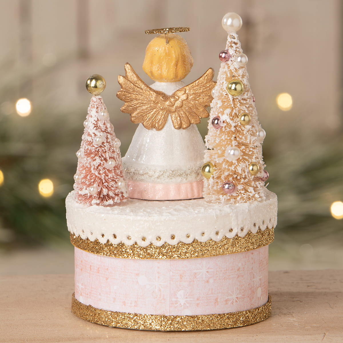 A delicate pink and gold bethany lowe angel figure with glittery wings on a festive box with miniature trees