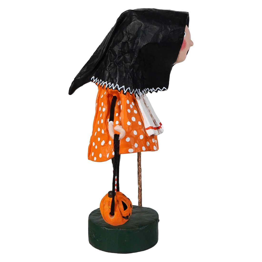 A lori mitchell halloween baba yaga figure in orange polka dot dress and black cloak holding a pumpkin lantern on green base