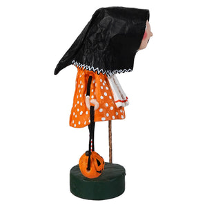A lori mitchell halloween baba yaga figure in orange polka dot dress and black cloak holding a pumpkin lantern on green base