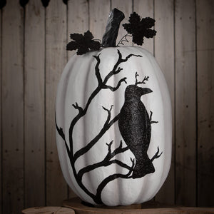 A white raven pumpkin decorated with a black glittery crow on bare branches with dark stem and leaf accents for halloween pumpkins and gourds