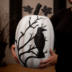 A white halloween pumpkin decorated with a black glittery raven perched on bare branches and glitter leaves