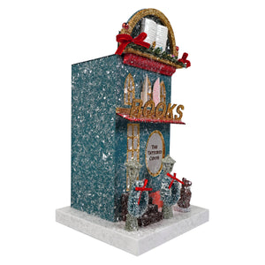 A glittery teal cody foster book shop figurine with gold trim and a red ribbon sign featuring miniature people in front