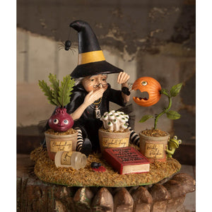 A botany witch figurine with a black and yellow hat and striped stockings sits with potted plants for halloween witches