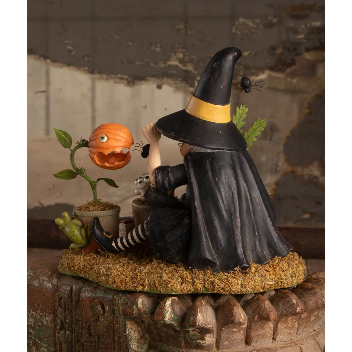 Black botany witch figurine with yellow striped hat and cape seated on mossy base next to pumpkin plant halloween witches