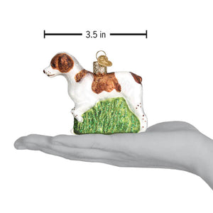 A white and brown glass brittany spaniel ornament with a glittery green grass base and a small golden hat on its back