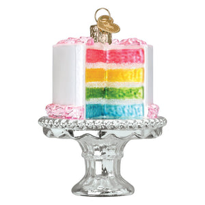 Colorful rainbow layered birthday cake ornament with pink frosting and gold heart tag on clear glass stand for dessert ornaments
