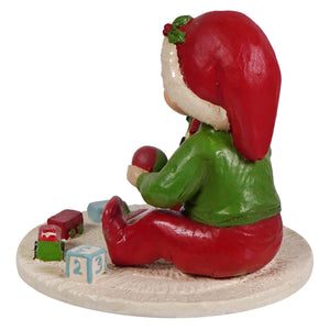 A glossy red and green elf figurine with a festive holly hat seated on a textured base with miniature toys from bethany lowe designs carson with train