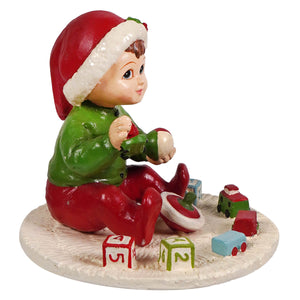 A glossy resin baby elf figurine in a red santa hat and green outfit sitting on snow with colorful blocks by bethany lowe designs
