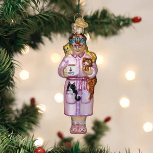 A pink glass christmas glass ornament features a cat lady ornament with a woman in pajamas holding a mug and two cats