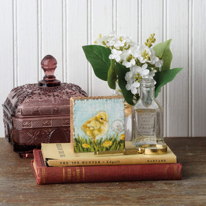 A vintage-style vignette featuring stacked books, a purple glass candy dish, white flowers in a bottle, and a decorative chick artwork.