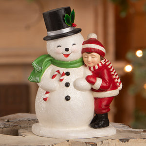 A cheerful child hugging a white bethany lowe designs snowman figurine with holly hat and candy cane details in winter attire