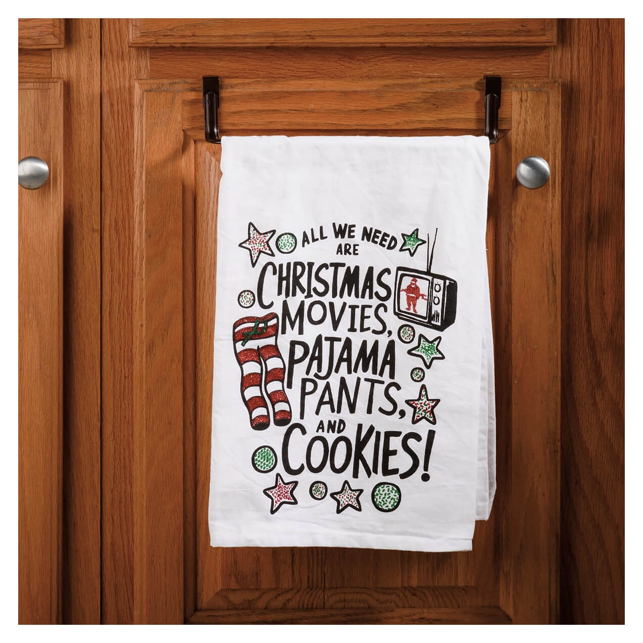 Decorative kitchen towel with festive Christmas-themed text and graphics hanging on a wooden cabinet door.