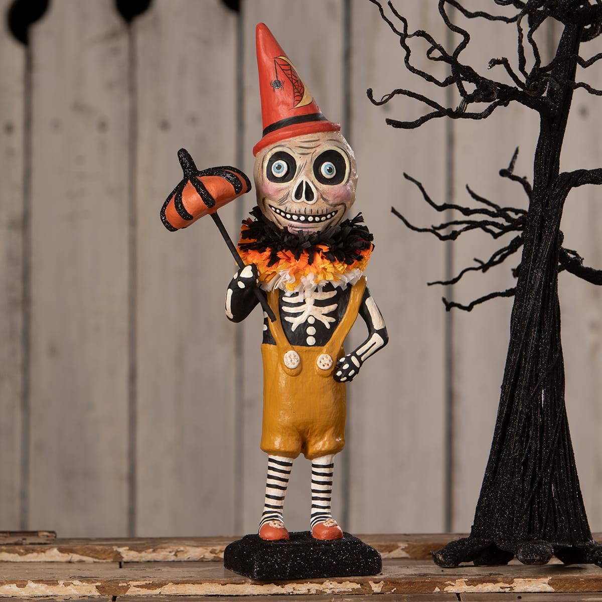 A whimsical orange and black skeleton decor figure dressed as a clown with a red spiderweb hat called circus skelly
