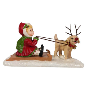 A festive bethany lowe figurine featuring a child in a red coat on a sled pulled by a tan puppy with antlers and a red nose