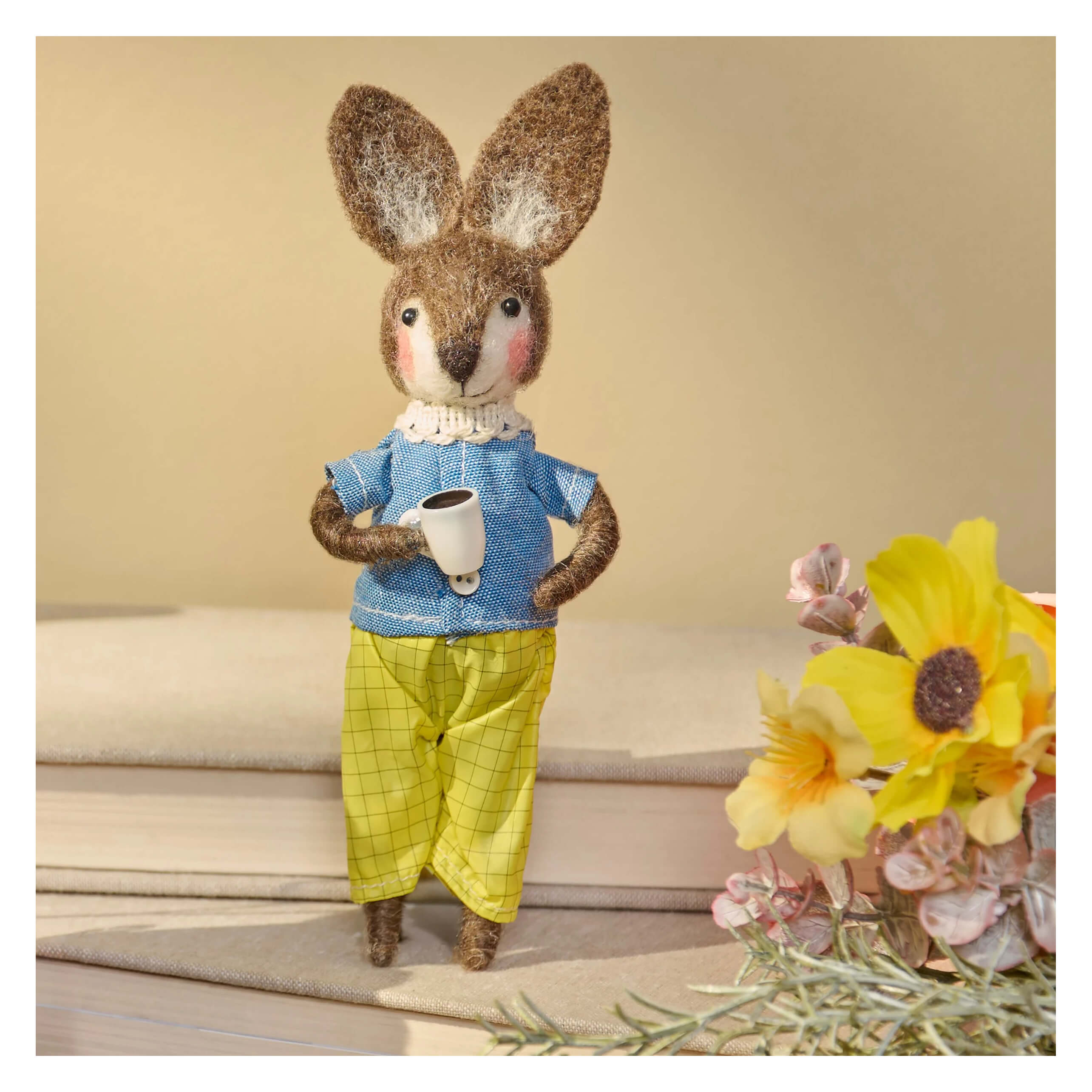 A brown felt coffee rabbit critter wearing a blue patterned shirt and yellow checkered pants holding a white mug
