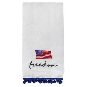 White tea towel with an embroidered American flag, ’freedom’ text, and blue pom-pom trim.