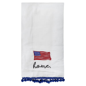 White tea towel with an embroidered American flag, ’home’ text, and blue pom-pom trim.