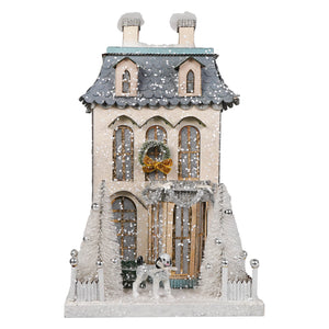 A festive white chateau house christmas village ornament with cream walls, gray roof, wreath, and frosted pine trees