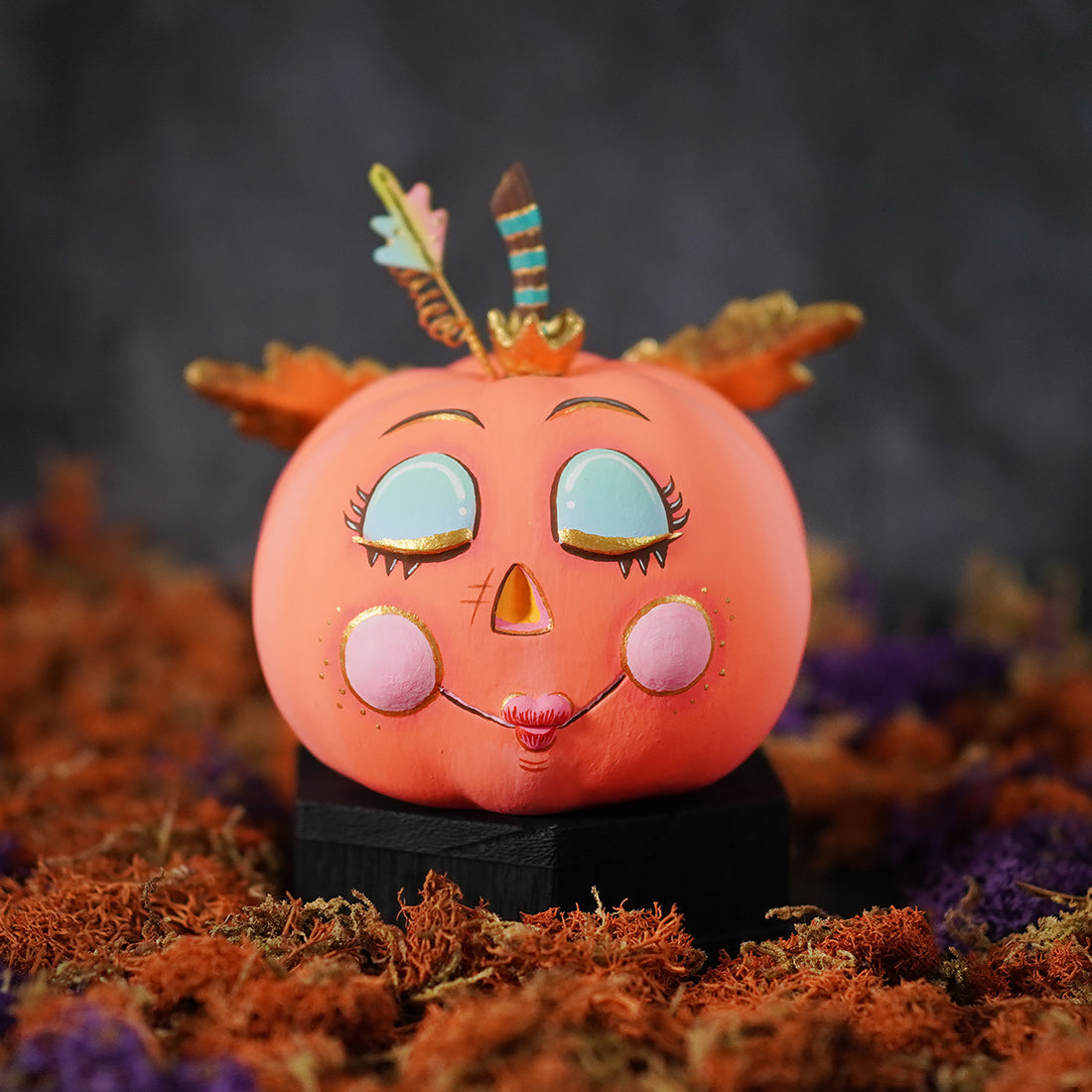 A whimsical demure peachy pink pixie pumpkin figurine with turquoise eyes pink cheeks and a feather crown perfect for pumpkin pixie display