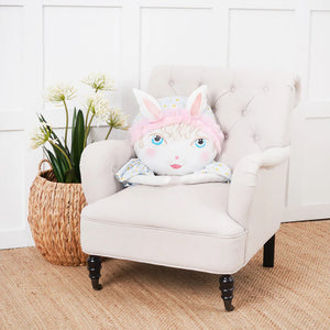 A plush pastel pink zombinaland decorative pillow with blue eyes and a soft white face on a tufted armchair